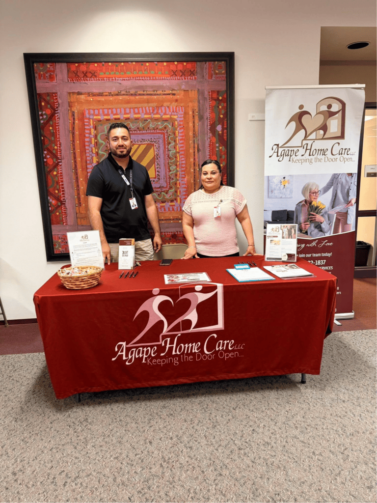 Two Agape Home Care representatives beside a branded information table and banner